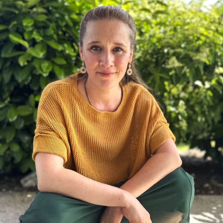 Rike Sylla Heilpraktikerin schaut in die Kamera, im Hintergrund ein grüner Busch.