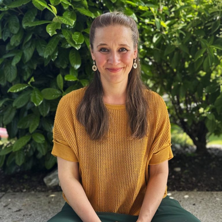 Rike Sylla, Heilpraktikerin lächelt in die Kamera, im Hintergrund ein grüner Busch.